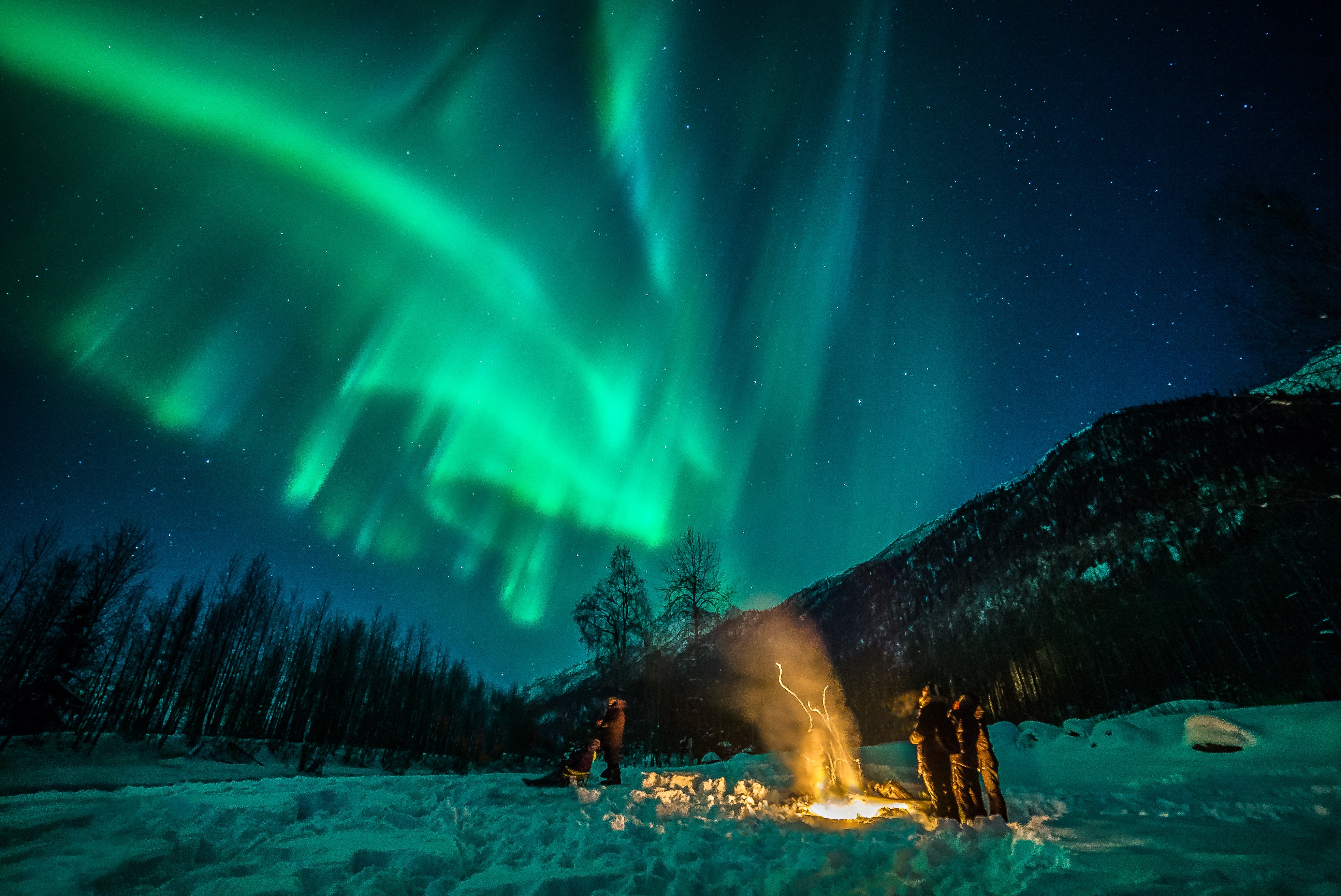 The best time to travel to Alaska: people around a fire watching the Northern Lights in the sky.